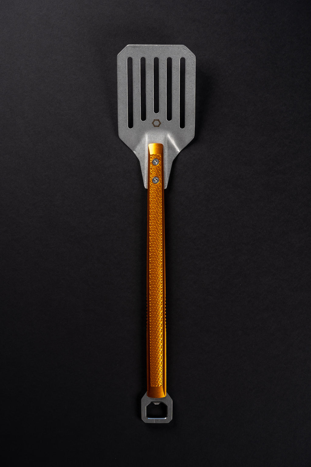 A metal kitchen spatula with a textured orange handle and a silver slotted head, shown vertically on a dark black background.
