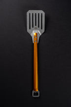 A metal kitchen spatula with a textured orange handle and a silver slotted head, shown vertically on a dark black background.