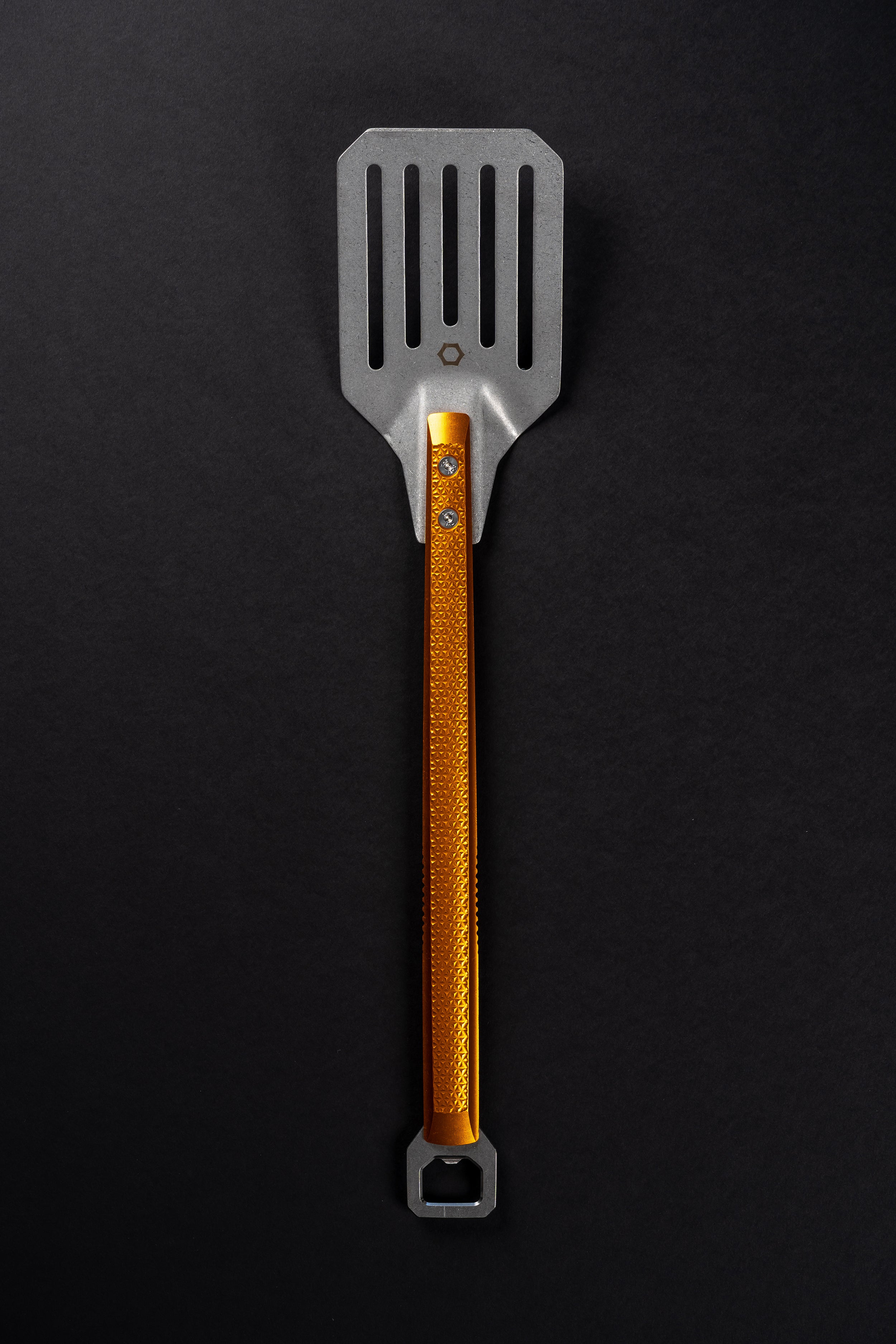 A metal kitchen spatula with a textured orange handle and a silver slotted head, shown vertically on a dark black background.