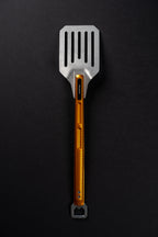 A metal kitchen spatula with a silver flat head and a textured orange handle, shown vertically on a dark black background.