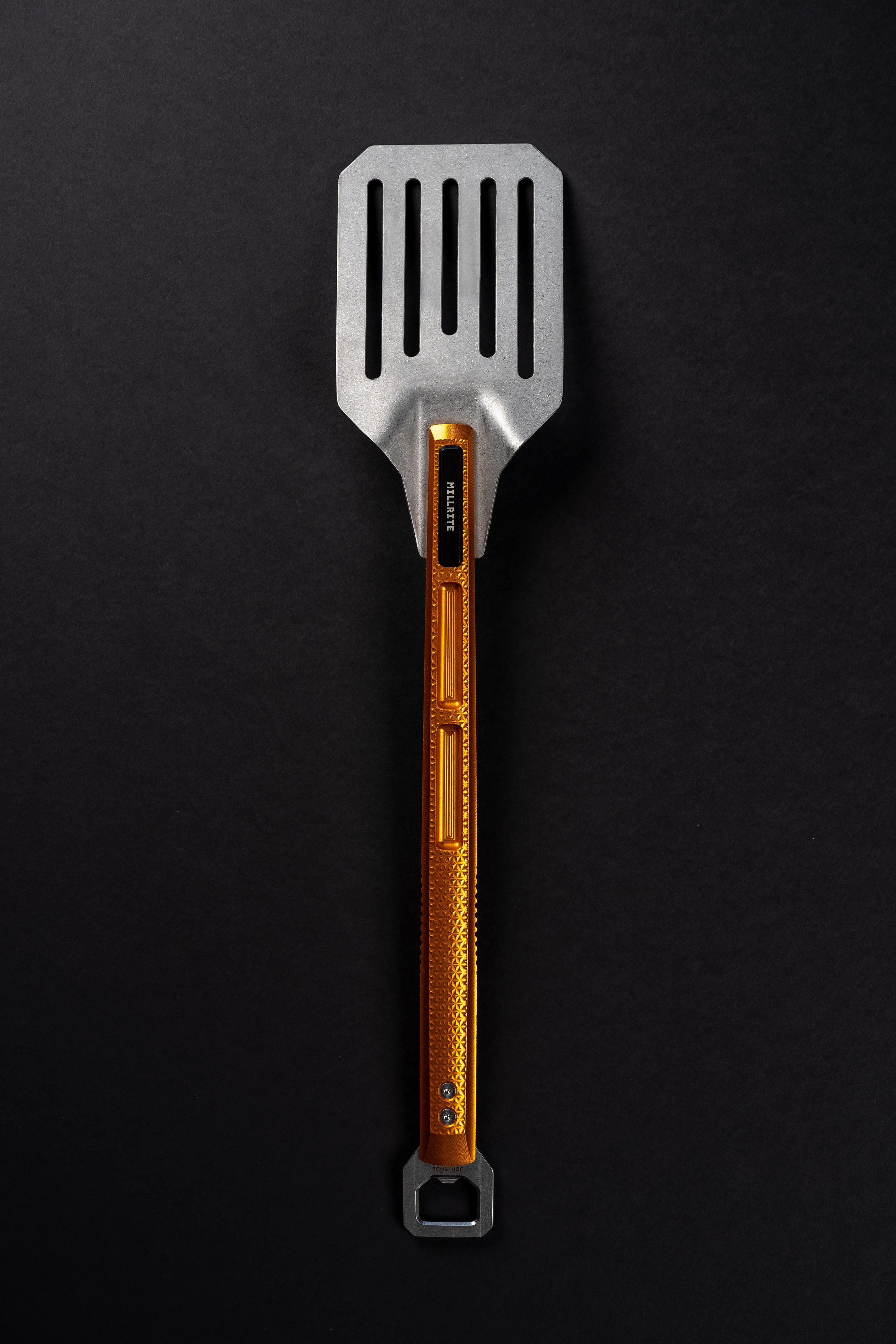 A metal kitchen spatula with a silver flat head and a textured orange handle, shown vertically on a dark black background.