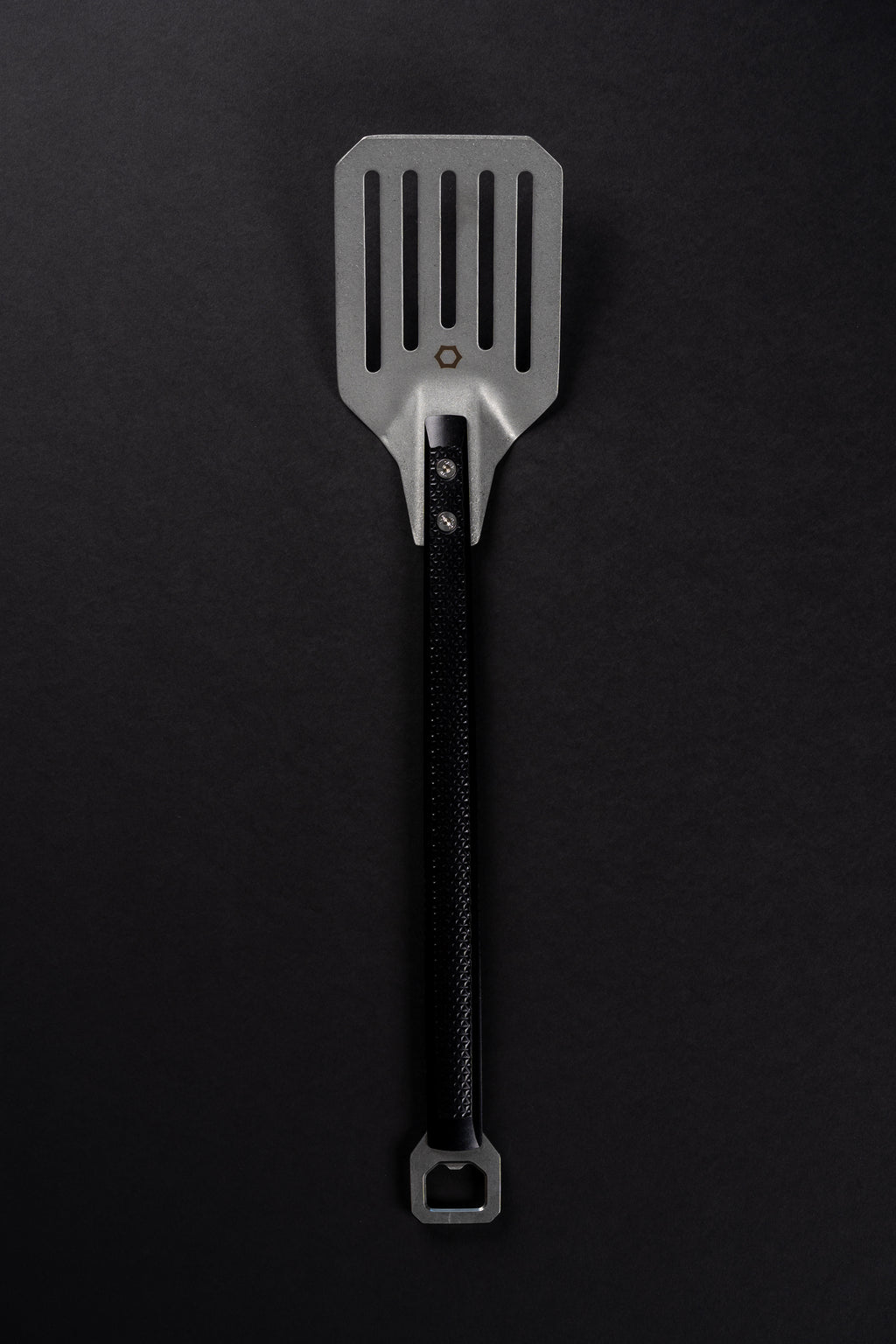 A metal spatula with a black machined handle, photographed vertically against a dark black background.