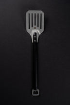 A metal spatula with a black machined handle, photographed vertically against a dark black background.