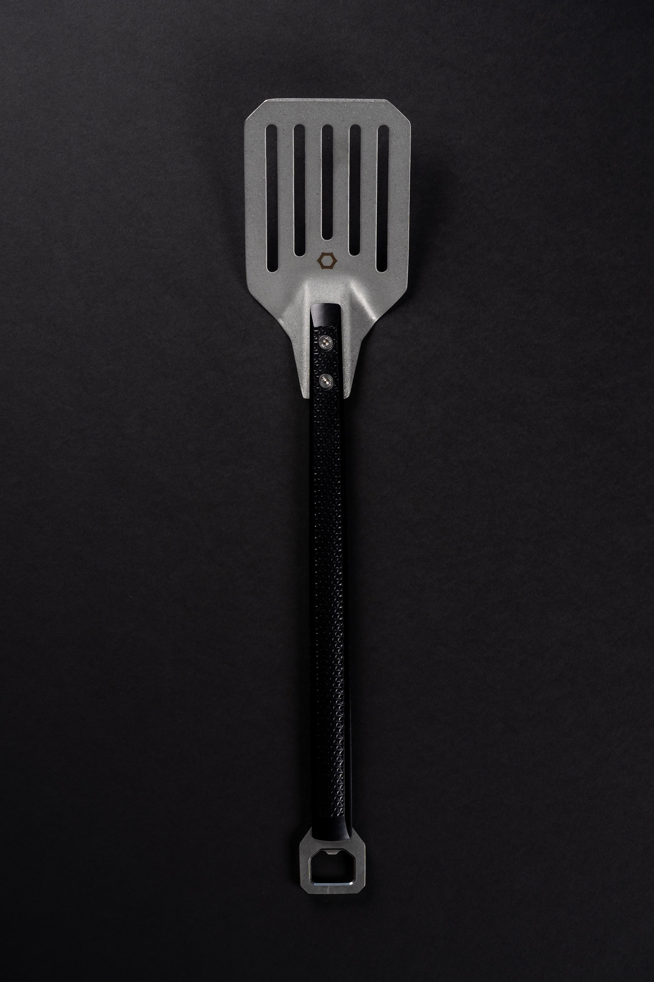 A metal spatula with a black machined handle, photographed vertically against a dark black background.