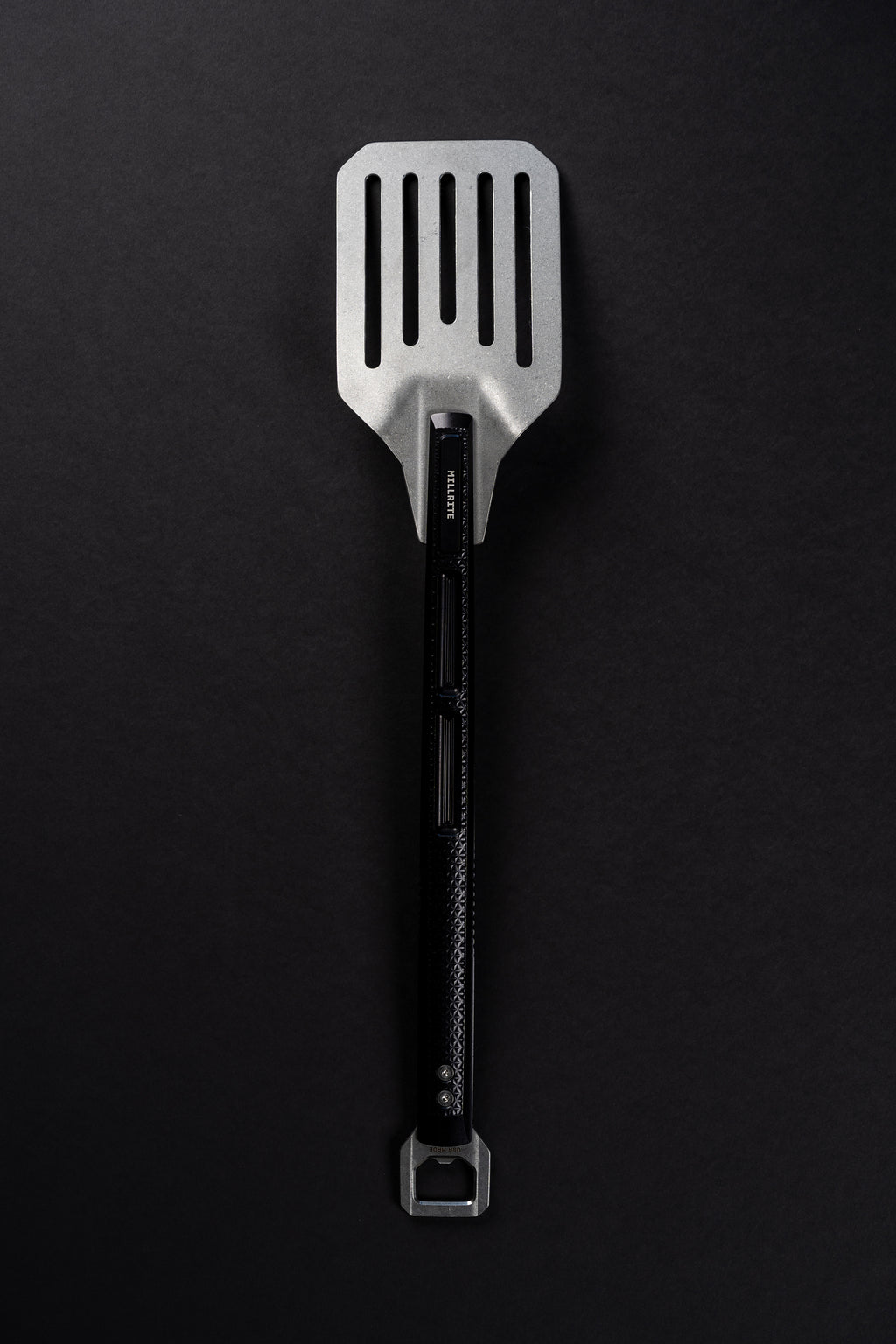 A stainless steel slotted spatula with a black handle and a bottle opener attachment at the end, photographed vertically against a dark textured background.