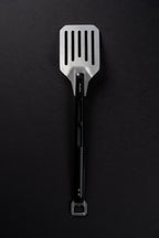 A stainless steel slotted spatula with a black handle and a bottle opener attachment at the end, photographed vertically against a dark textured background.
