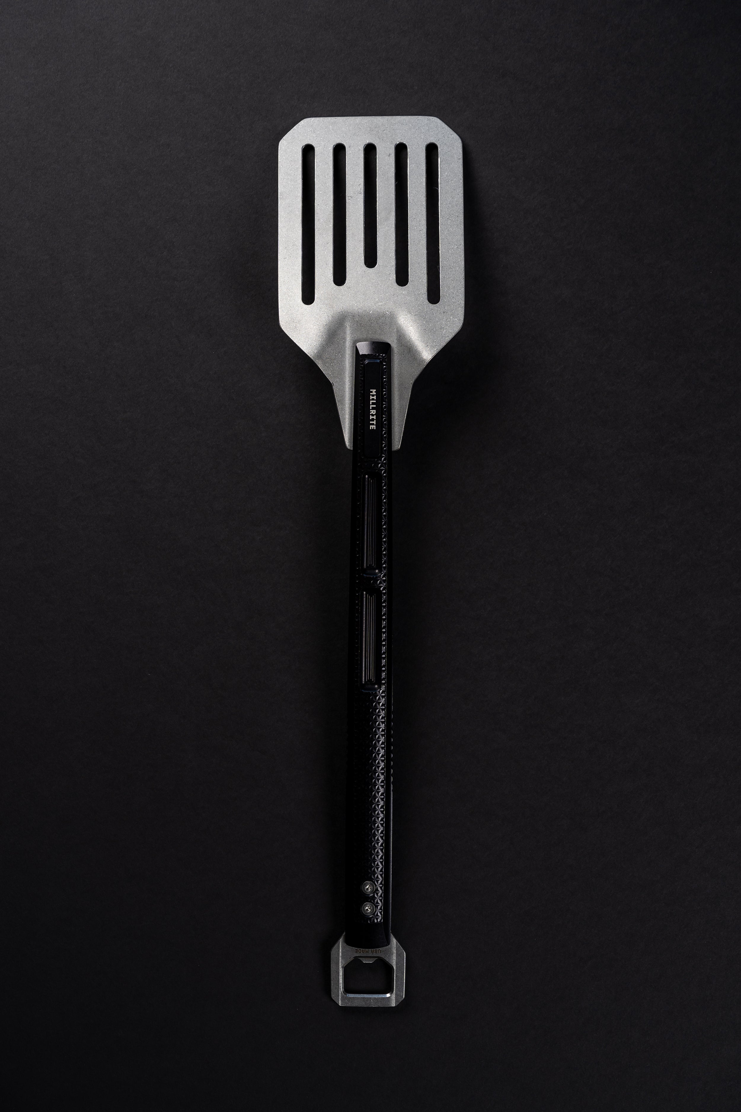 A stainless steel slotted spatula with a black handle and a bottle opener attachment at the end, photographed vertically against a dark textured background.
