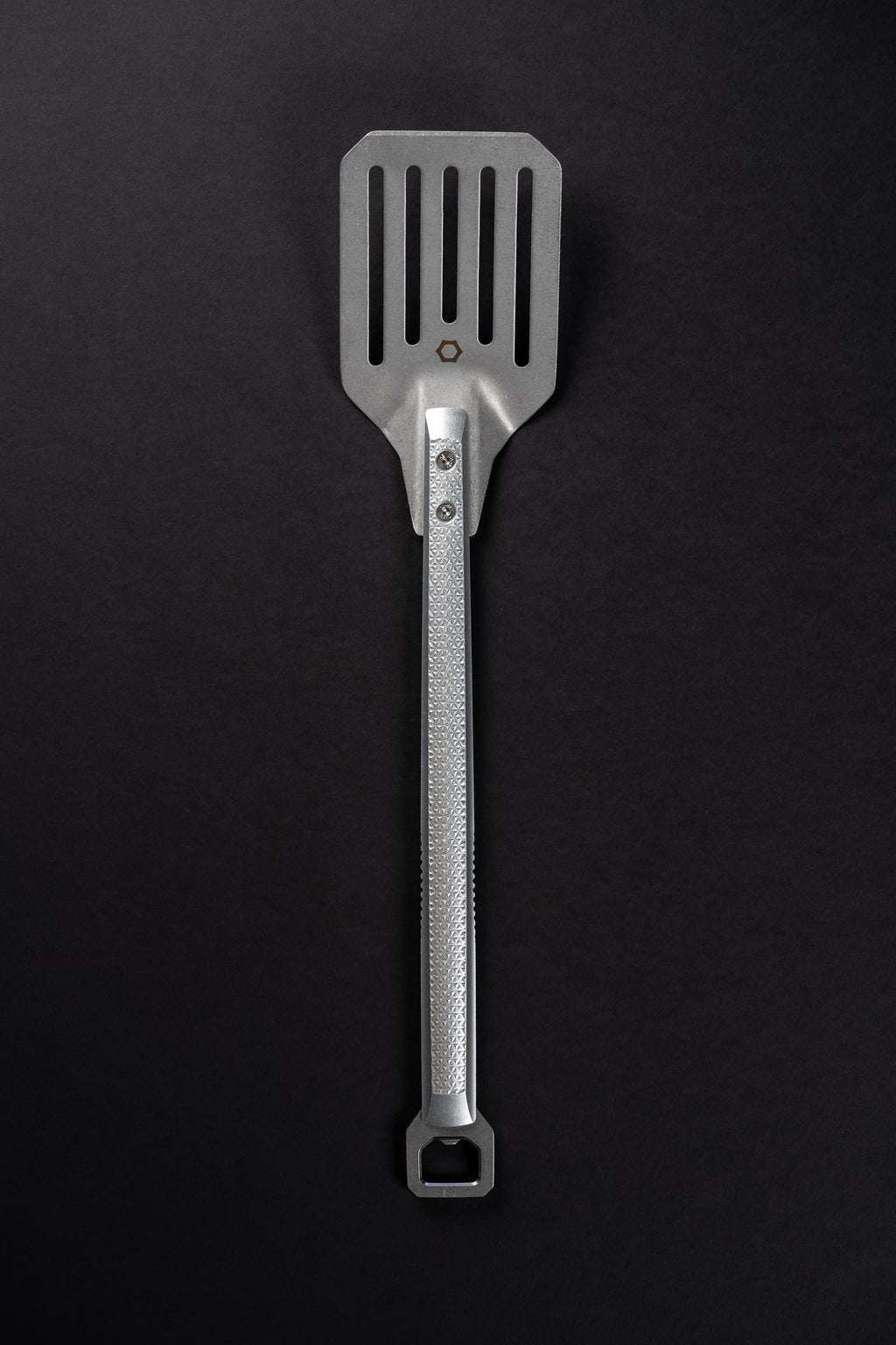 A stainless steel kitchen spatula with a textured handle and a slotted blade, shown vertically against a dark black background.