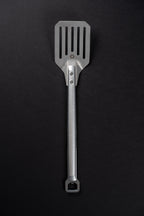 A stainless steel kitchen spatula with a textured handle and a slotted blade, shown vertically against a dark black background.