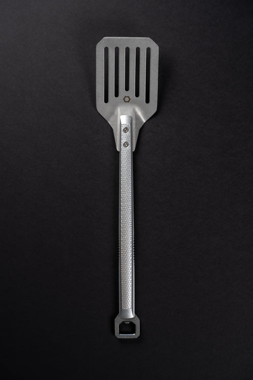 A stainless steel kitchen spatula with a textured handle and a slotted blade, shown vertically against a dark black background.