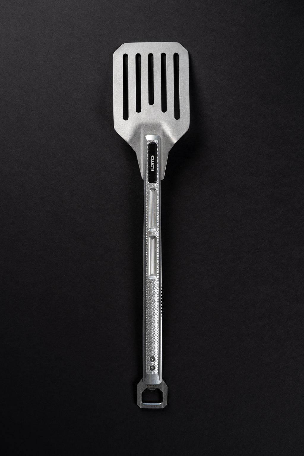 A stainless steel slotted spatula with a textured handle and a hanging loop at the end, photographed vertically against a dark textured background.