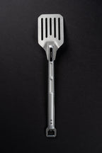 A stainless steel slotted spatula with a textured handle and a hanging loop at the end, photographed vertically against a dark textured background.