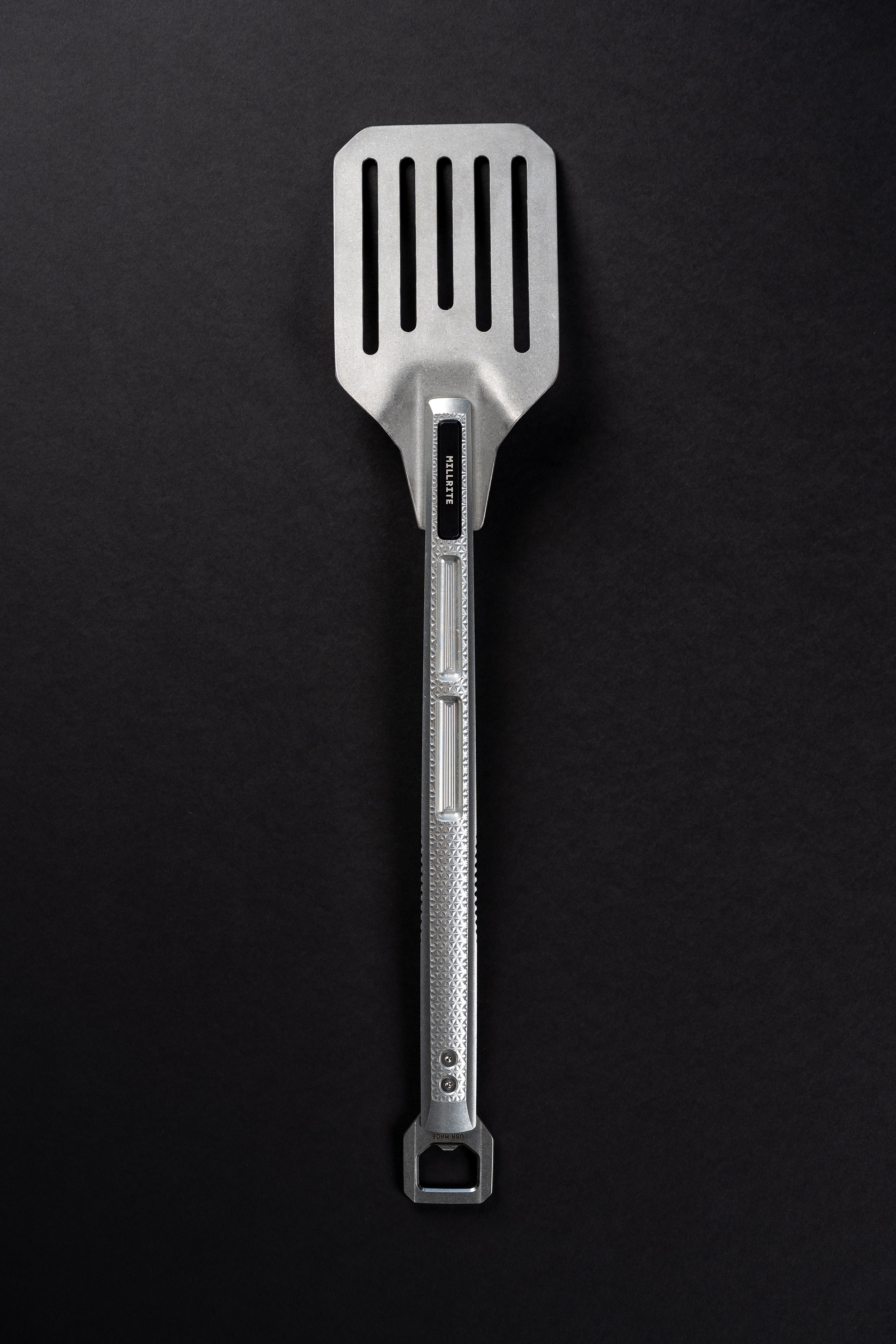 A stainless steel slotted spatula with a textured handle and a hanging loop at the end, photographed vertically against a dark textured background.