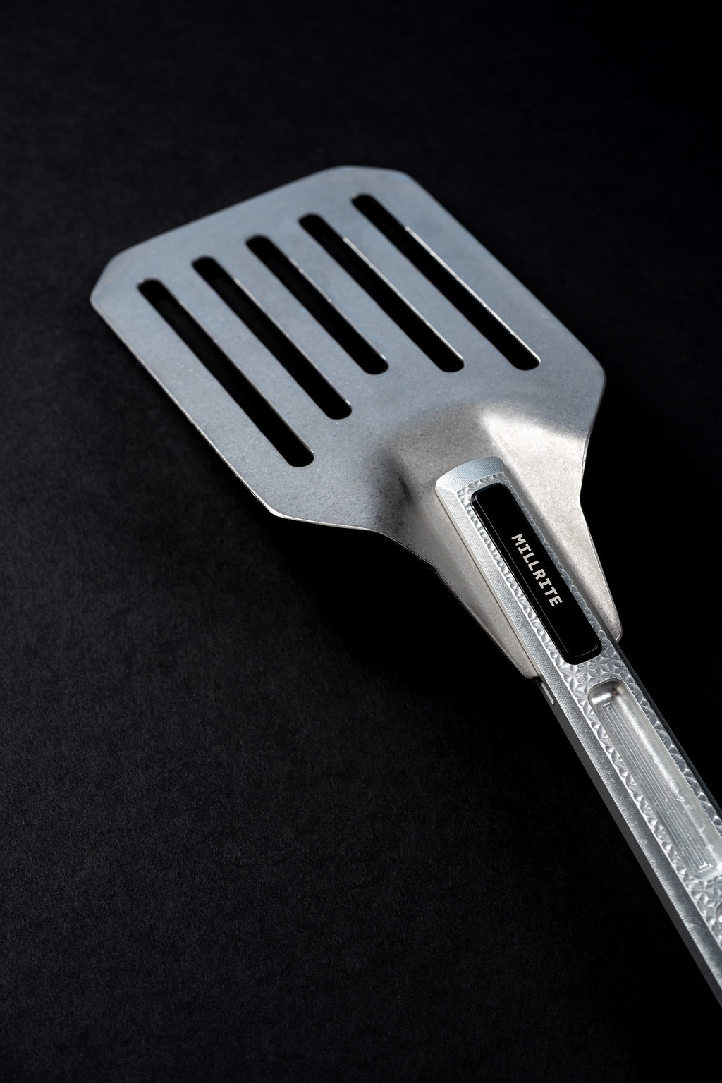 A stainless steel slotted spatula with a black handle grip and the brand name 'MILLRITE' laser etched on it, set against a solid black background.