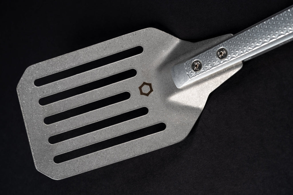 A close-up of a metallic gray spatula with a textured handle, shown against a solid black background.