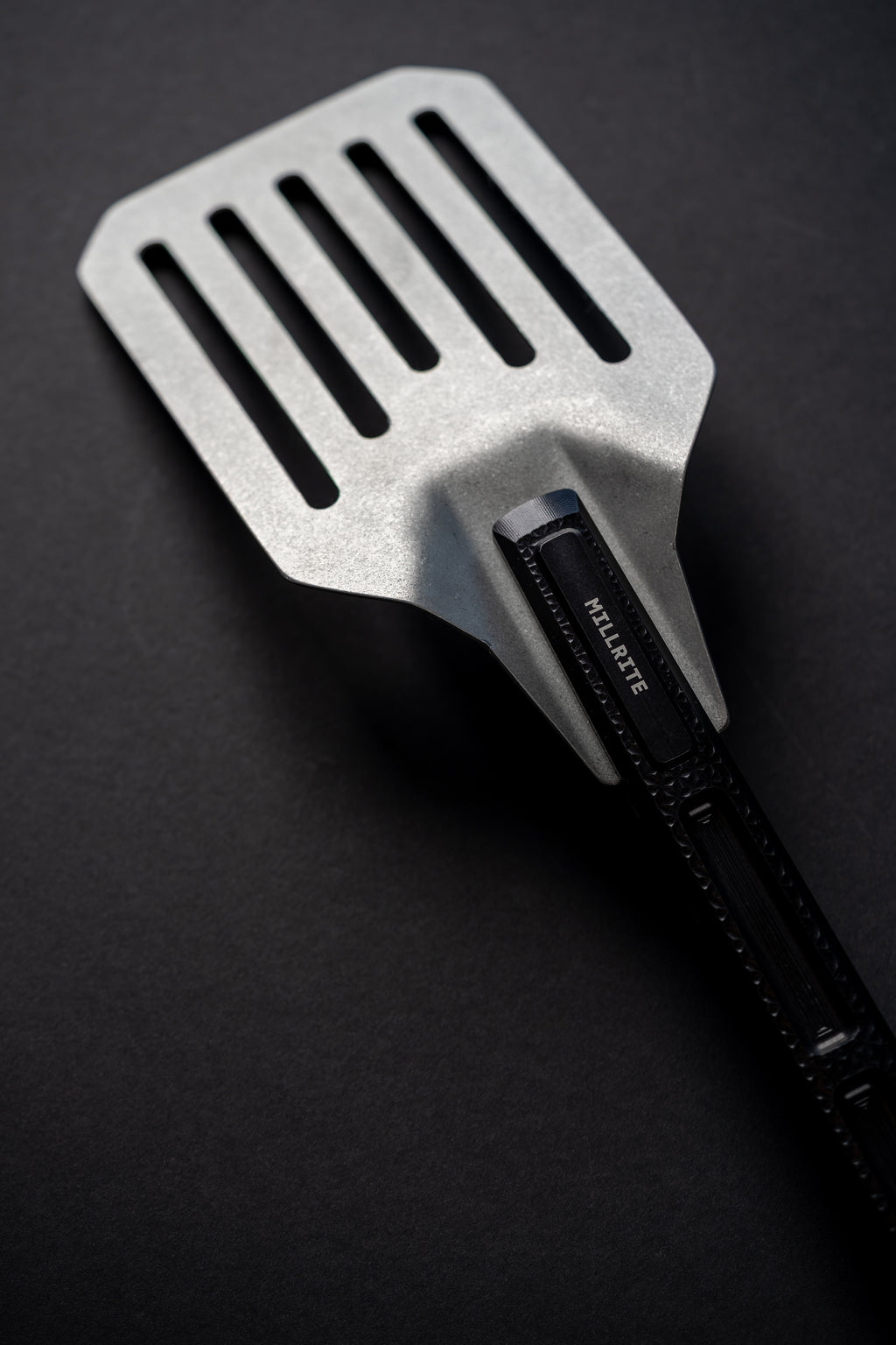 A metal spatula with a black handle, photographed diagonally on a dark textured background. The brand name 'MILLRITE' is clearly printed on the handle.
