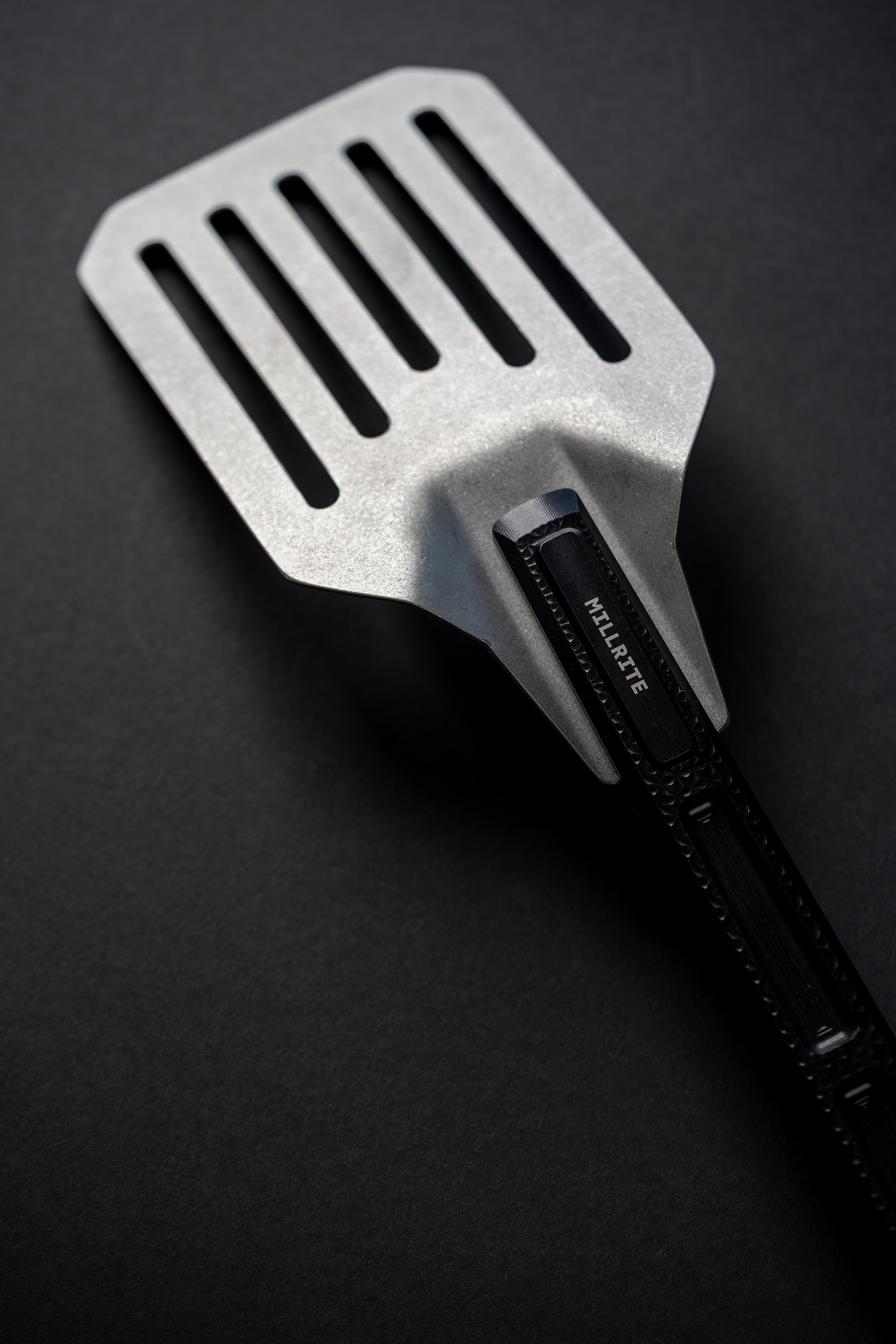 A metal spatula with a black handle, photographed diagonally on a dark textured background. The brand name 'MILLRITE' is clearly printed on the handle.