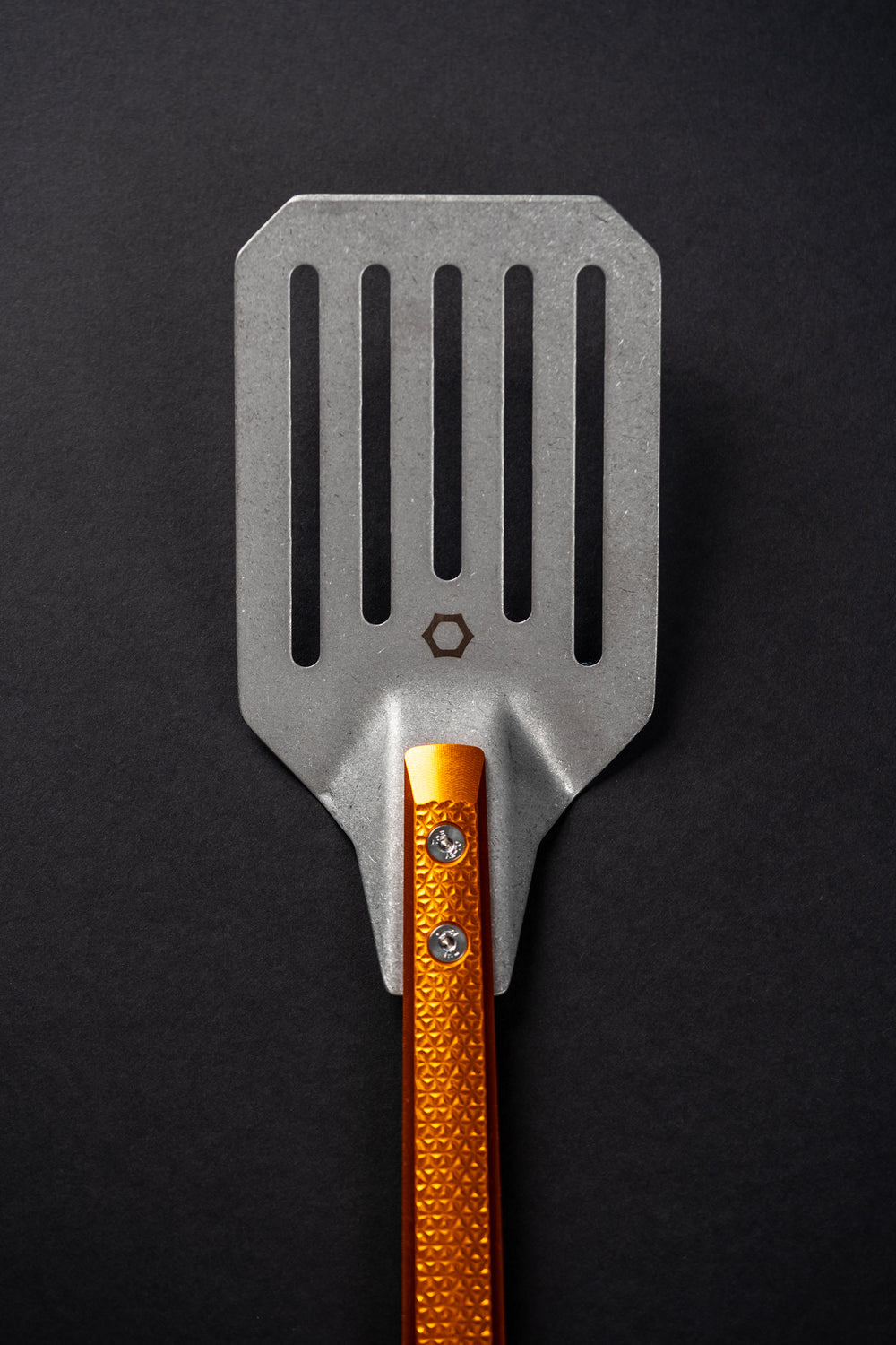 A metal grill spatula with a textured orange handle, photographed vertically against a dark black background. The spatula head is made of silver-toned metal with vertical slots and a small hexagonal logo near the center.