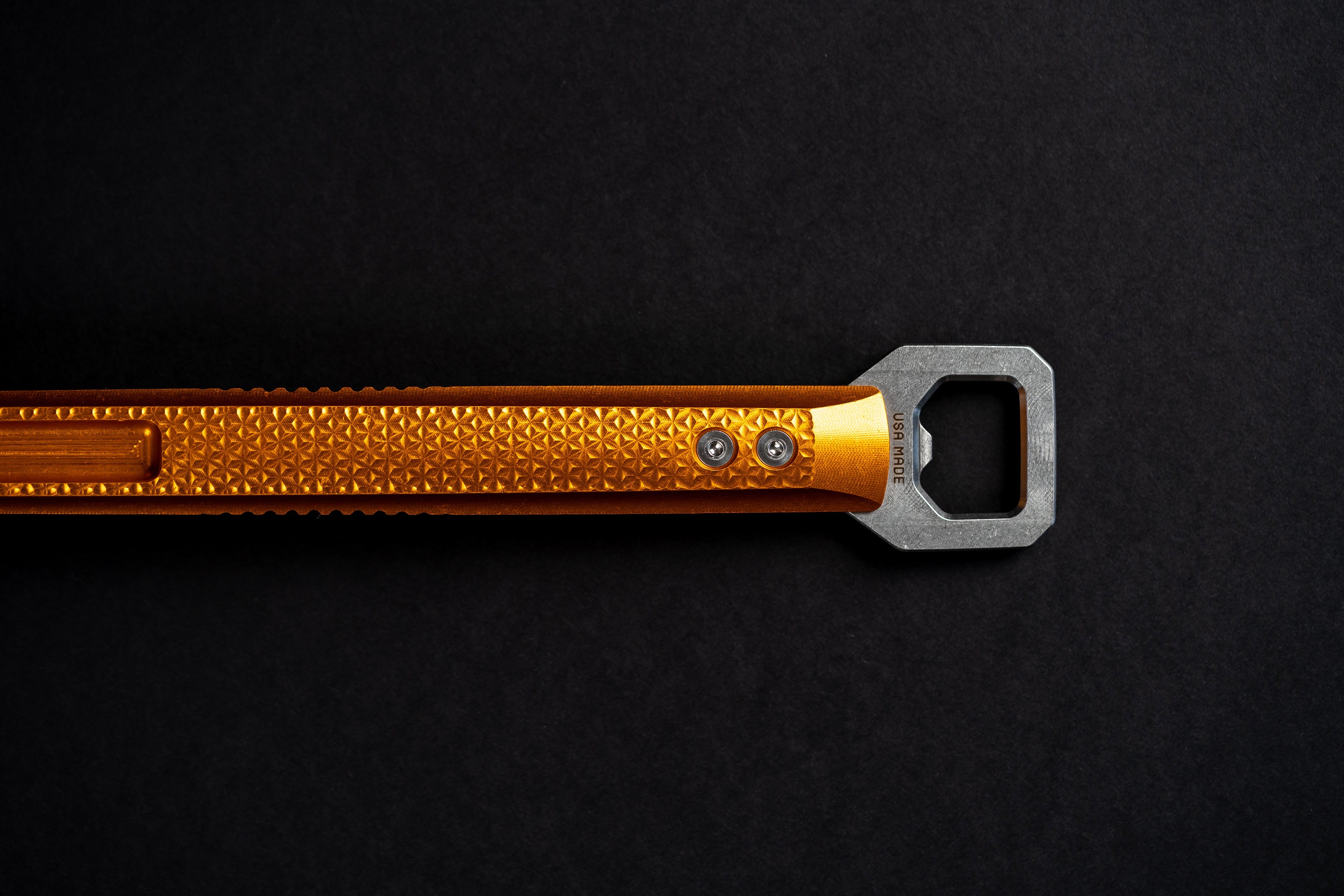 A close-up of a metallic orange bottle opener with a textured handle and a silver metal blade, positioned horizontally on a dark black background.