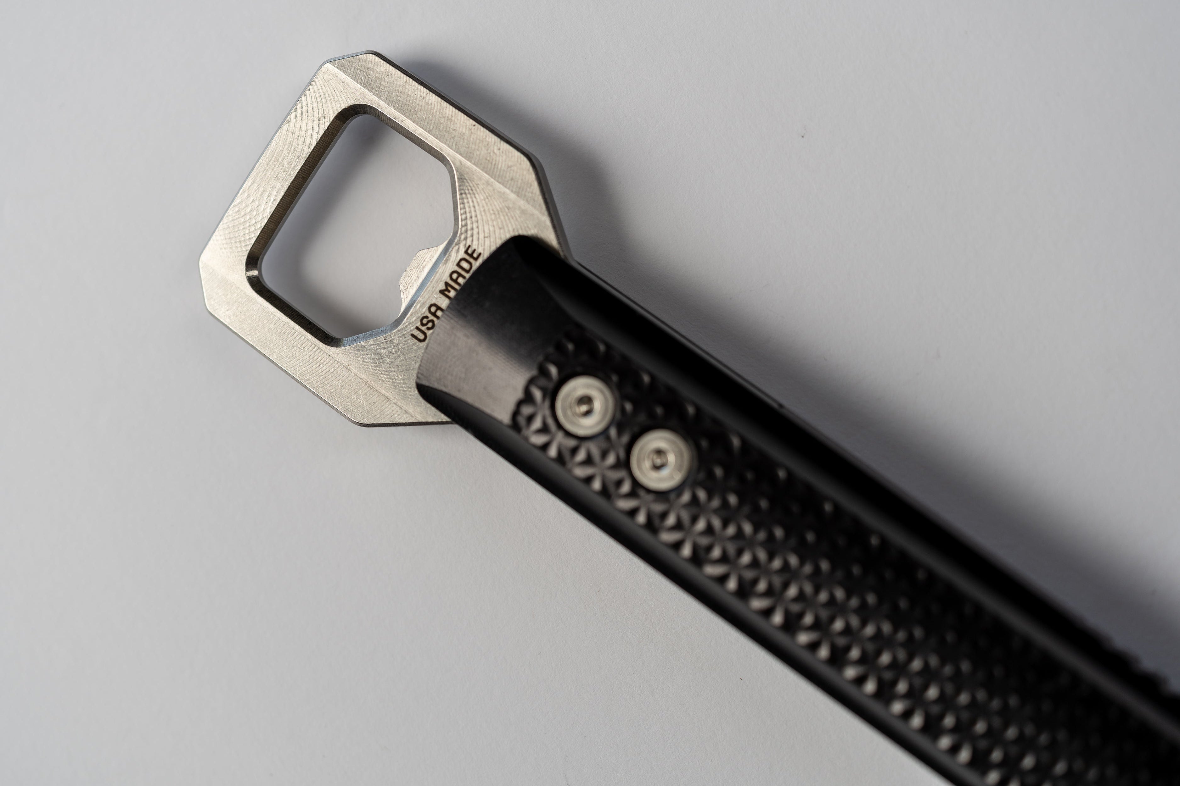 A close-up of a bottle opener with a silver metallic geometric blade and a black textured handle, photographed on a light gray background.